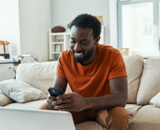 Person seated on a couch using a smartphone with an open laptop on their lap, in a well-lit living room