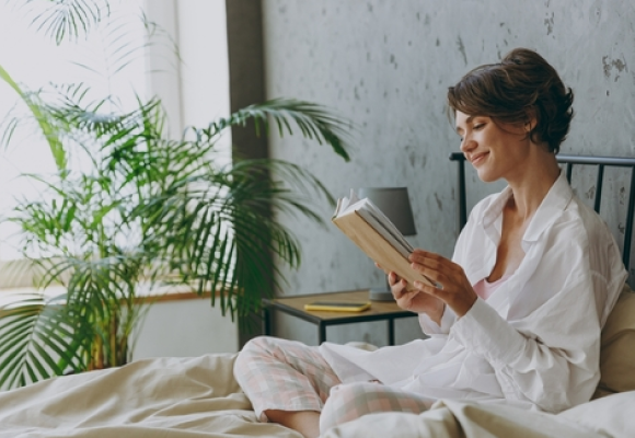 Young calm happy woman wear white shirt pajama and lying in bed read book study rest relax spend time in bedroom lounge home