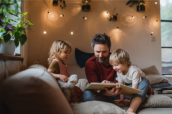 Father reading to children in cozy living room with warm lighting and indoor plants